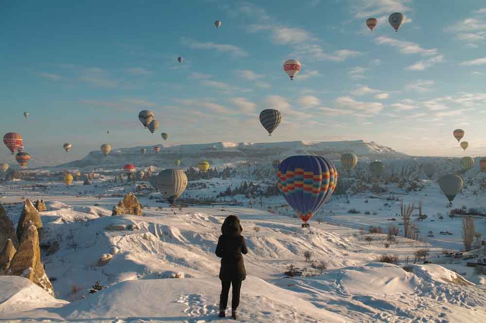 Person standing on snowy hill, watching colorful hot air balloons float above a winter landscape under a blue sky in Cappadocia, Turkey. The scene conveys wonder and serenity.