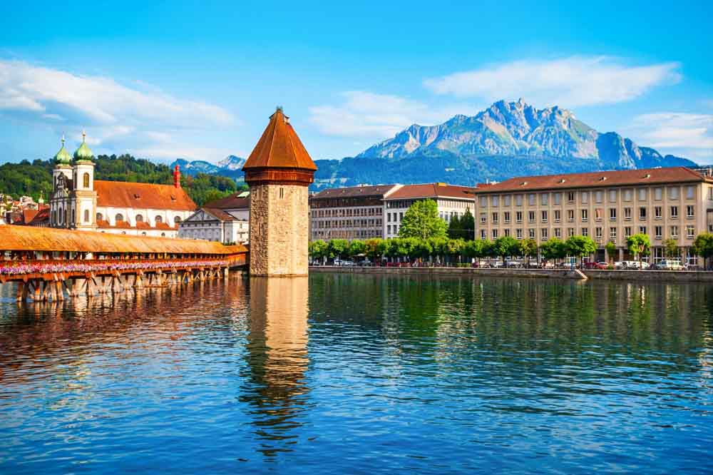 Beautiful view of Chapel Bridge (Kapellbrücke) in Lucerne, Switzerland, with its iconic water tower, historic buildings, and the majestic Mount Pilatus in the background — a must-visit destination on Switzerland tour packages.