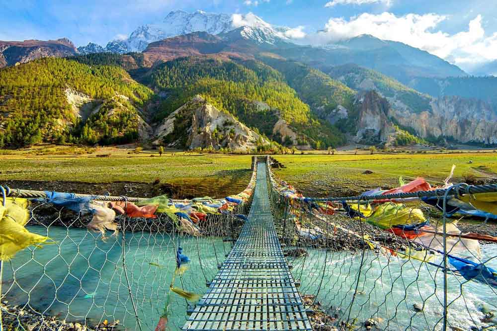 A suspension bridge with colorful prayer flags crosses a clear blue river, leading to a lush, tree-covered valley with snow-capped mountains in the background.