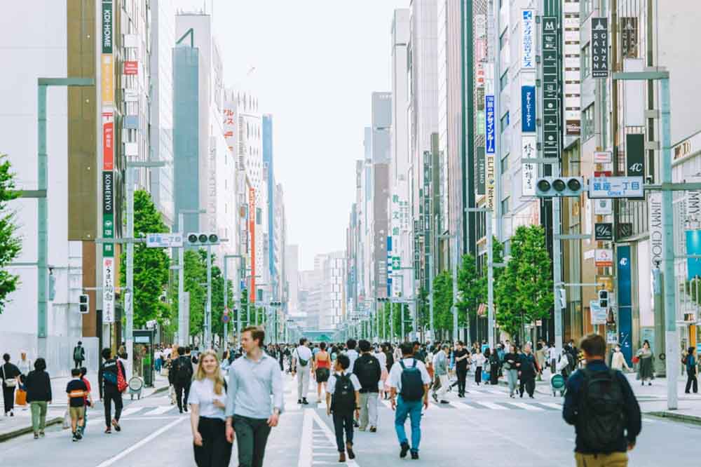 Bustling city street scene in daylight, with diverse pedestrians walking among tall buildings featuring vibrant signs. The atmosphere is lively and urban.
