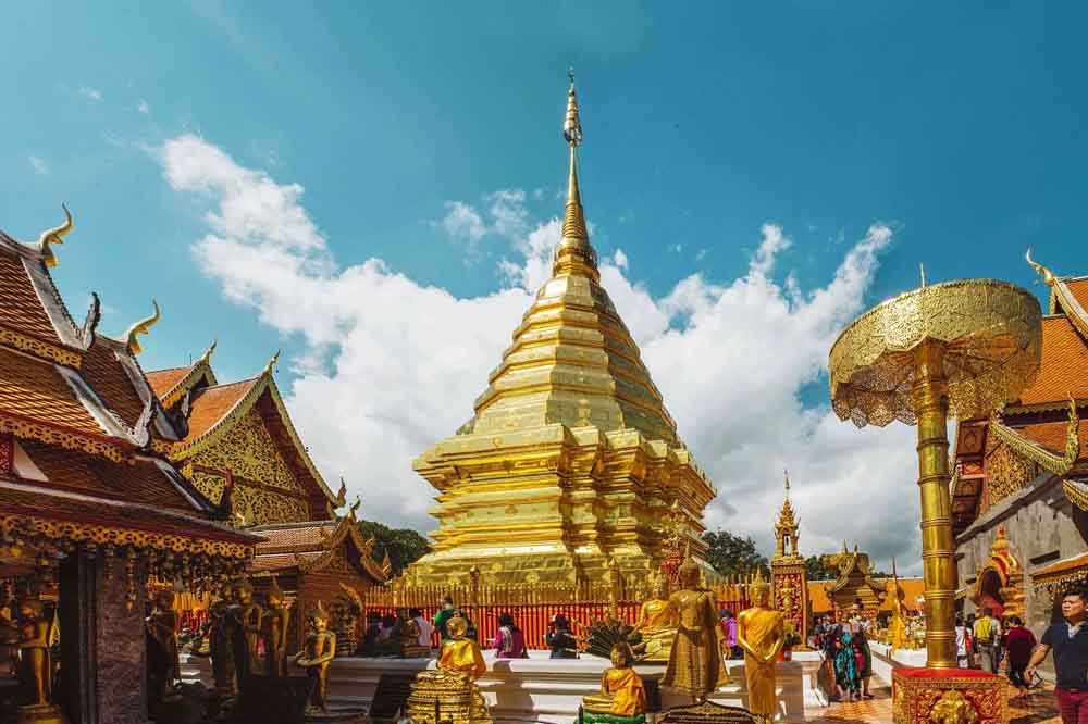 Golden temple spire under a vibrant blue sky with fluffy clouds, surrounded by ornate red and gold buildings and statues. Visitors wander, creating a lively atmosphere.