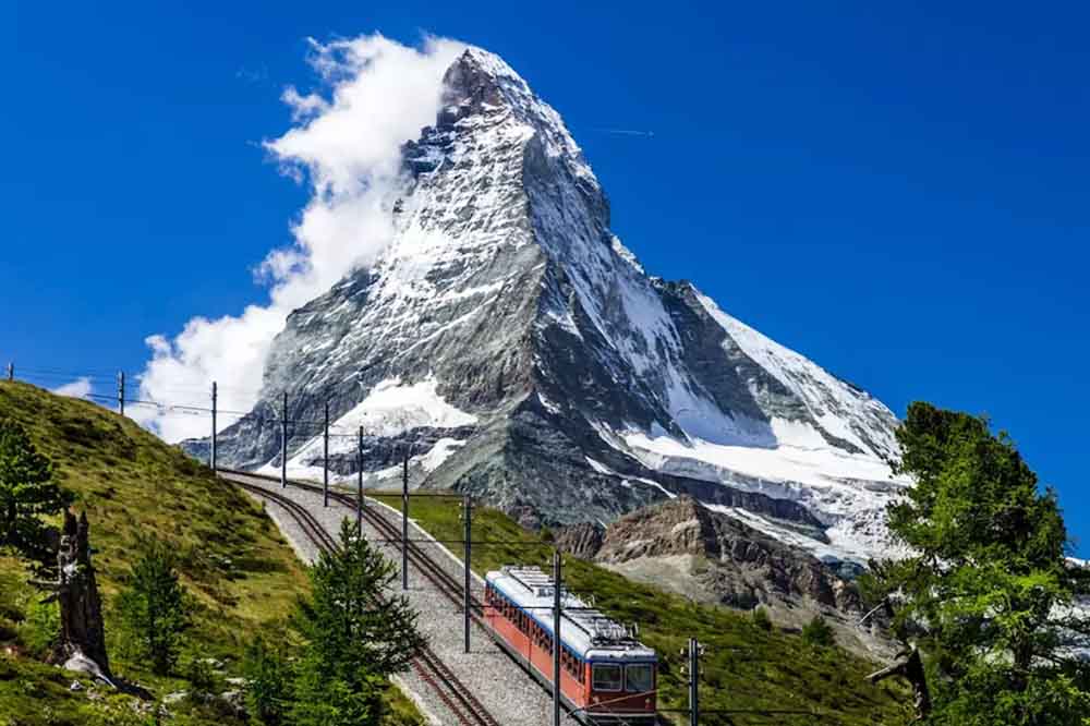 A train travels on tracks with the snow-capped Matterhorn mountain in the background, under a clear blue sky. Greenery surrounds the scene.