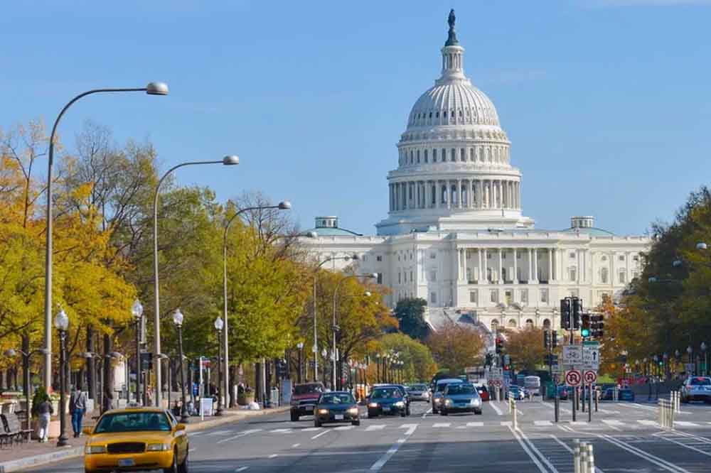 Wide view of the U.S. Capitol under a blue sky, flanked by autumn trees and a busy street with cars and a yellow taxi in Washington, D.C.