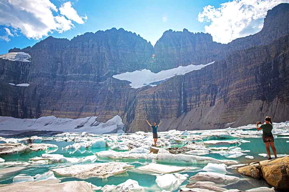 A person stands on icy rocks with arms raised at a glacial lake. Majestic cliffs and blue sky set a dramatic, awe-inspiring scene. Another person takes a photo.
