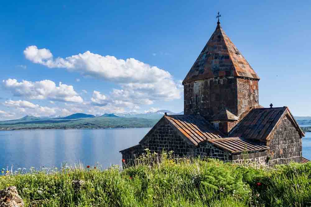 Ancient stone church with a pointed roof stands amid green grass, overlooking a serene lake under a bright blue sky with fluffy clouds. Peaceful atmosphere.