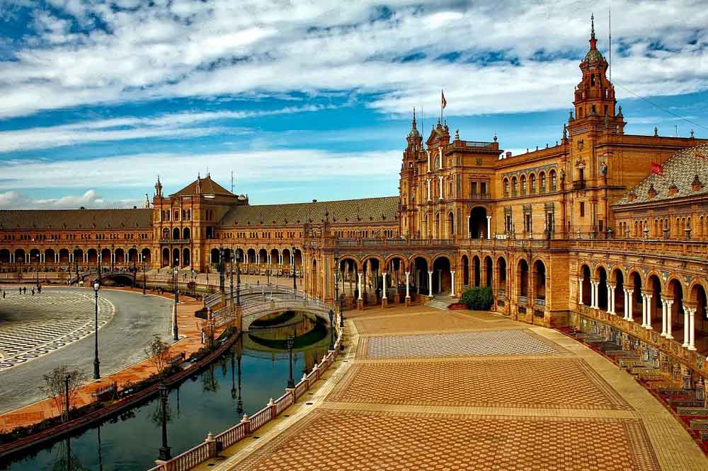 Historic plaza on a sunny day, featuring a curved brick building with arches and towers. A reflecting canal and patterned walkways enhance the grandeur.