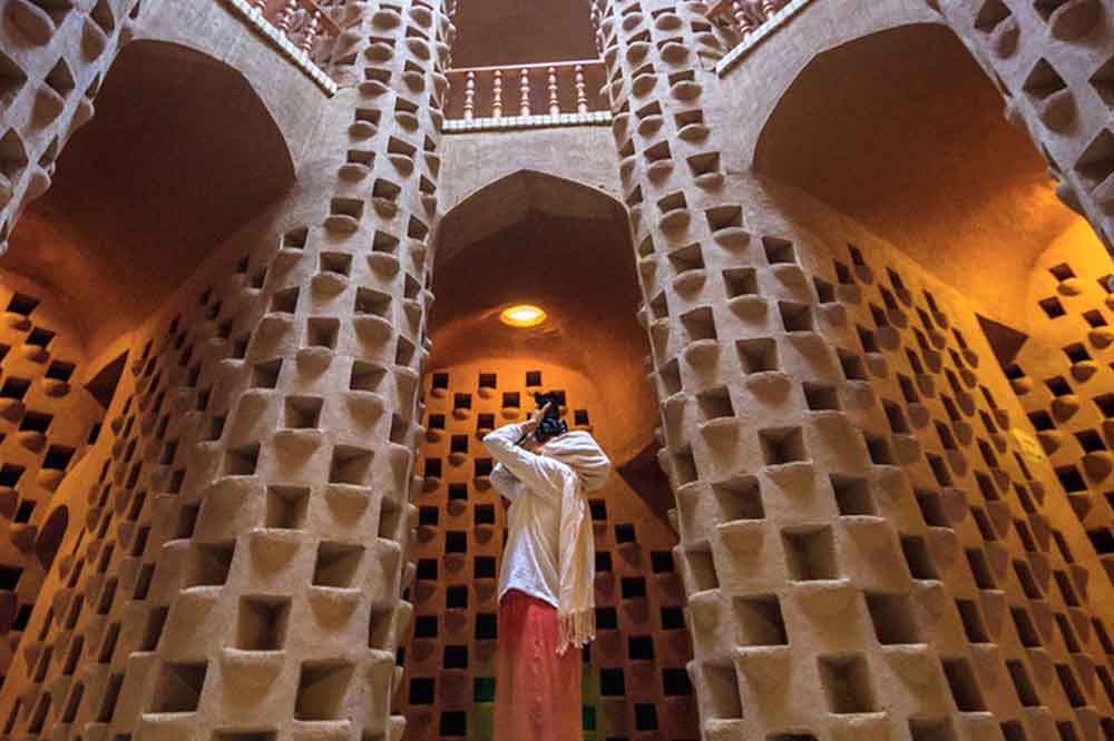 A person in colorful attire photographs upward in a vast, intricately designed clay structure with geometric patterns and arched ceilings. The scene conveys awe.