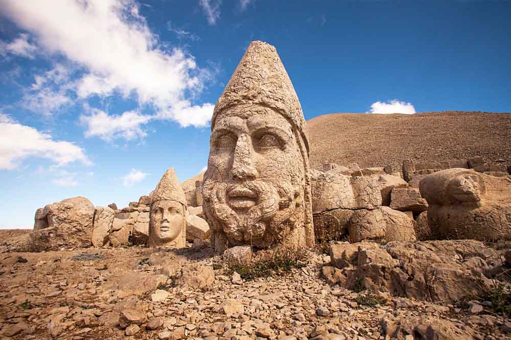 Ancient stone heads on rocky terrain at Mount Nemrut, Turkey, under a vibrant blue sky with light clouds. The scene feels timeless and monumental.