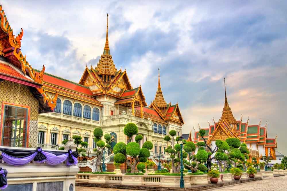 Ornate Thai palace with intricate golden roofs, colorful facades, and manicured gardens, set against a vibrant sky at sunset. Majestic and serene atmosphere.