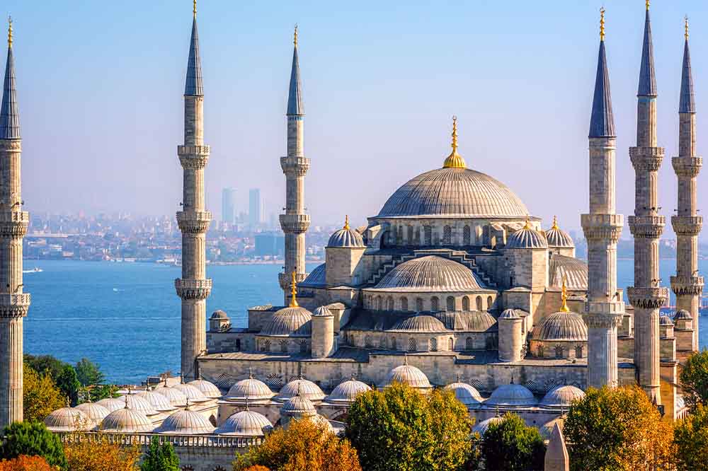A majestic mosque with six tall minarets and a large central dome overlooks a tranquil blue sea. Autumn trees frame the foreground, adding warmth.