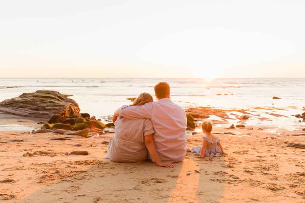A family of three sits on a sandy beach at sunset. The parents sit closely, embracing, while the child plays nearby. Calm ocean waves and rocks in the background.