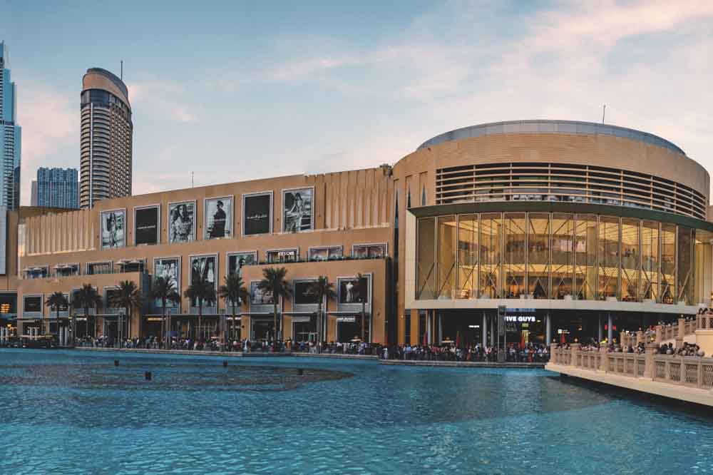 A modern, lit-up shopping mall by waterfront at dusk, featuring palm trees and adjacent skyscrapers. The scene is lively, with a clear evening sky.