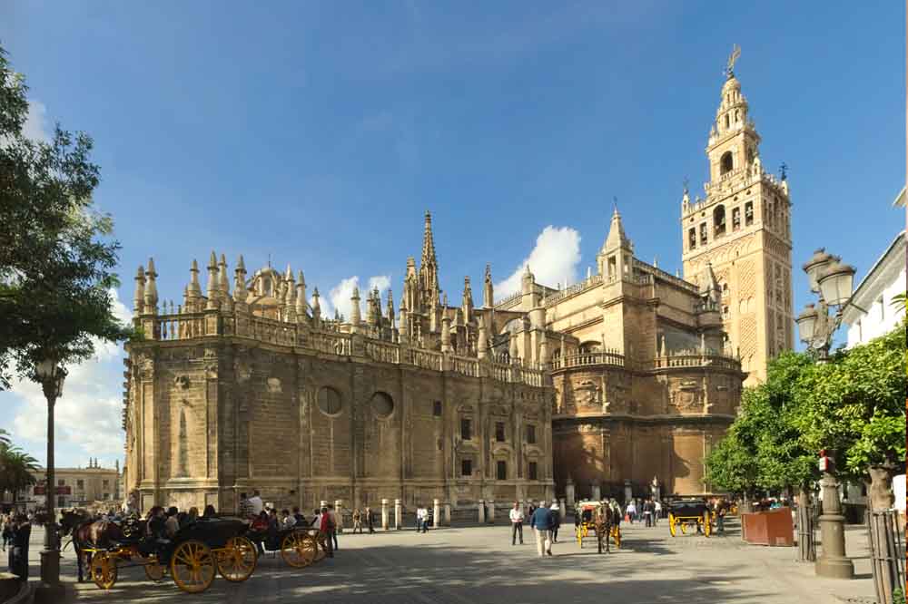 A historic cathedral under a clear blue sky, with a tall bell tower and ornate stonework. Horse-drawn carriages and people walk in the sunny plaza.