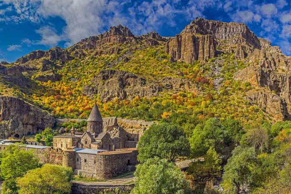 Historic monastery nestled in a lush valley, surrounded by vibrant autumn foliage and majestic rocky mountains under a vivid blue sky. Peaceful ambiance.