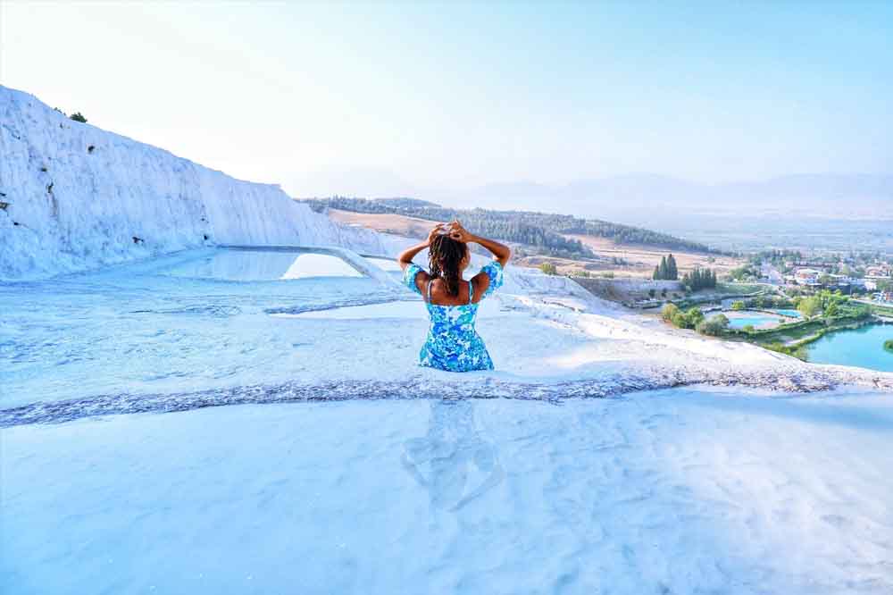 Woman in a blue dress stands in shallow, iridescent pools of Pamukkale, Turkey. Stepped white terraces stretch towards a hazy landscape and town below.
