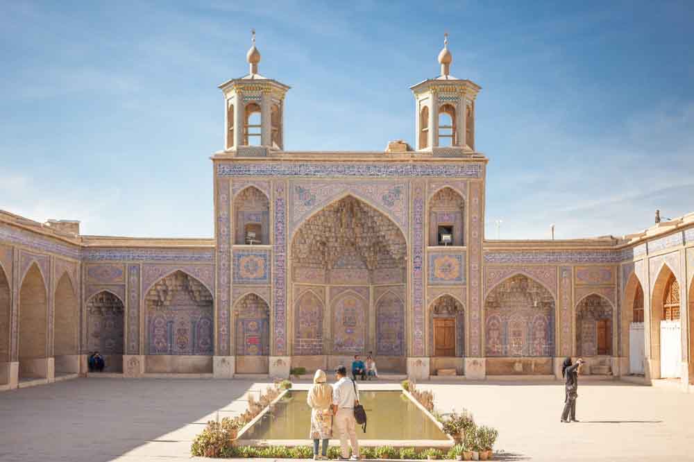 Historic mosque courtyard with intricate tilework, central pool, and two towers under a blue sky. Visitors admire and photograph the serene architecture.