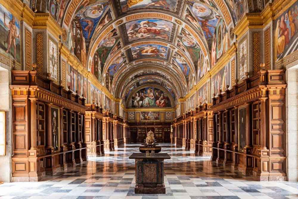 Ornate library hall with a vaulted ceiling richly adorned with colorful frescoes. Elegant wooden shelves line the walls, creating a grand and historic ambiance.