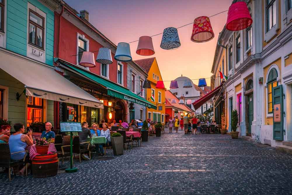 Colorful street scene at dusk with vibrant buildings and people dining outdoors. Hanging lampshades adorn the street, creating a cozy, lively ambiance.