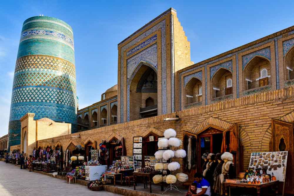 A vibrant market scene in Khiva, Uzbekistan, features a towering, intricately tiled turquoise minaret beside a historic brick madrasa. Stalls display colorful goods along the street.