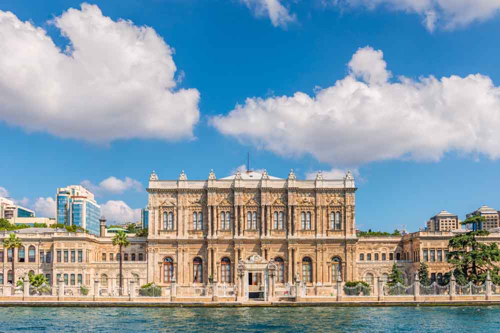Historic palace facade with ornate architecture by the water, under a bright blue sky with fluffy clouds, conveying grandeur and serenity.