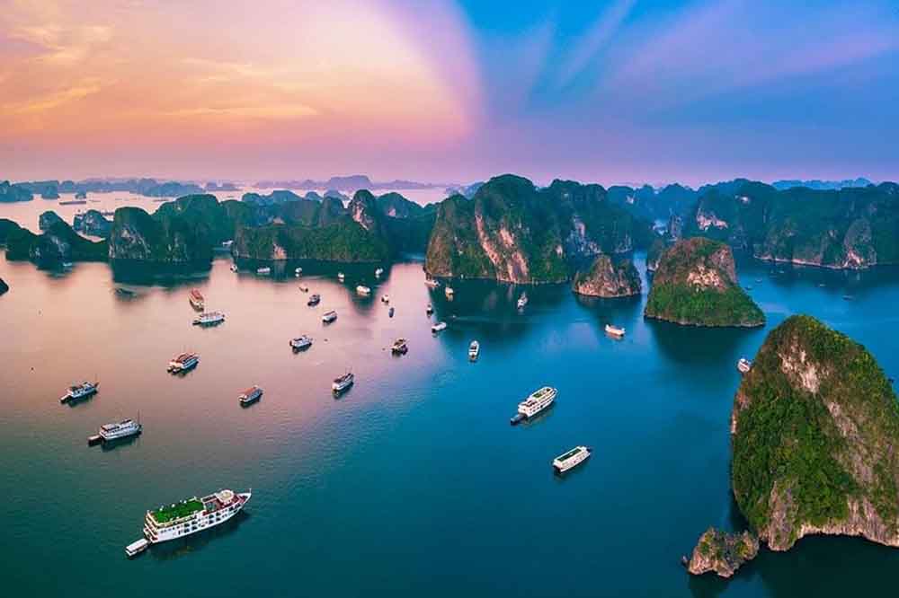 A picturesque aerial view of numerous boats sailing in a serene blue sea surrounded by lush, rocky islands under a vibrant, colorful sunset sky.