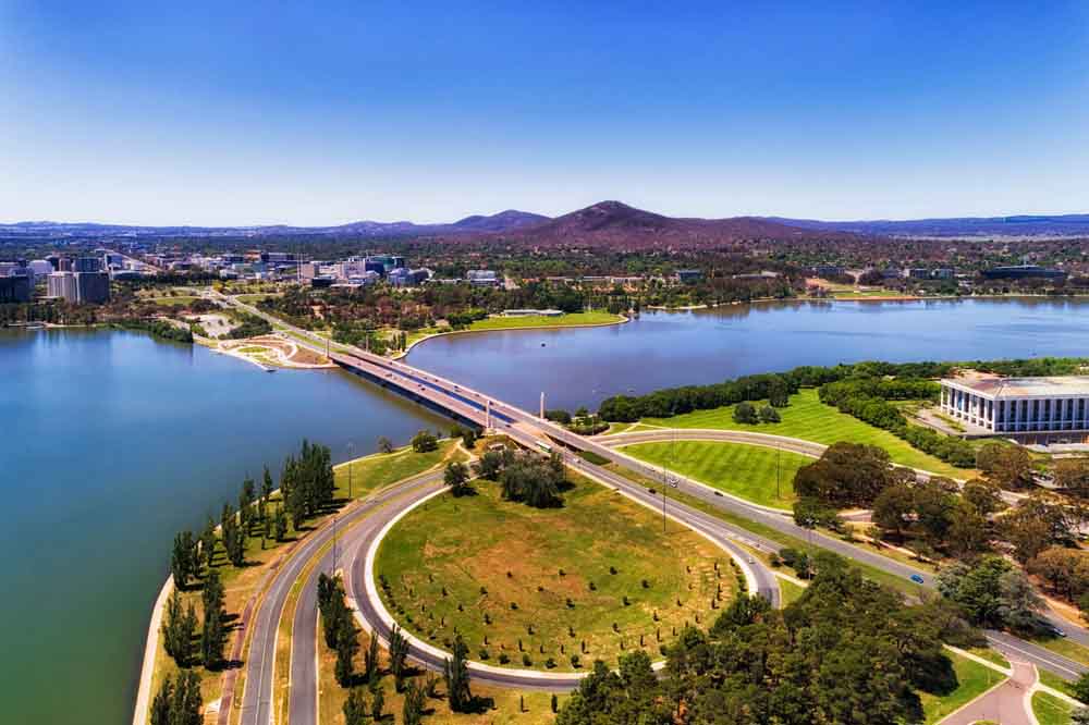 Aerial view of a scenic cityscape featuring a curved road, lush greenery, a river, and a distant city skyline under a clear blue sky. Calm and vibrant.