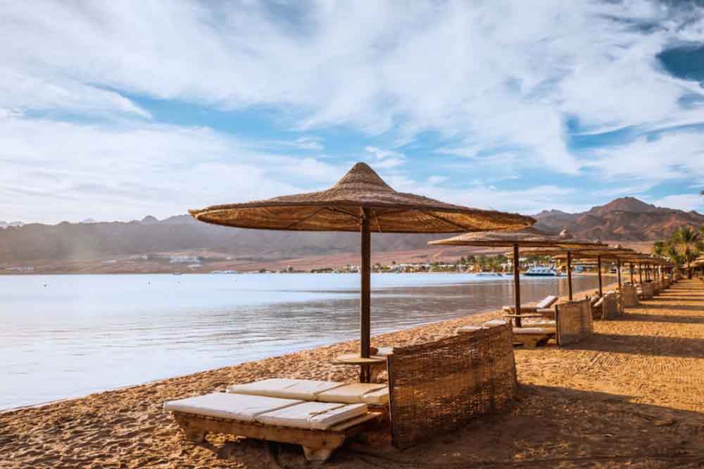 Sandy beach with rows of empty sun loungers under straw umbrellas facing a calm, blue sea. Mountains and fluffy clouds create a serene backdrop.