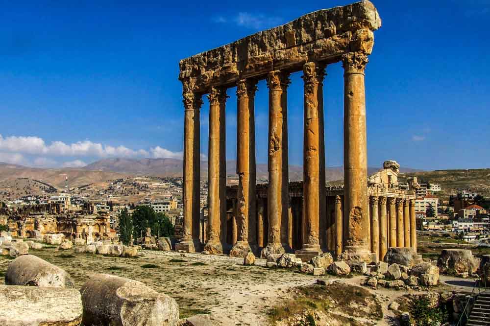 Ancient Roman temple ruins with tall, weathered columns under a clear blue sky. Surrounding landscape features distant mountains and a small town.