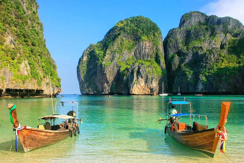 Two wooden boats float on clear turquoise water, surrounded by towering, lush green cliffs under a bright blue sky. The scene is peaceful and idyllic.