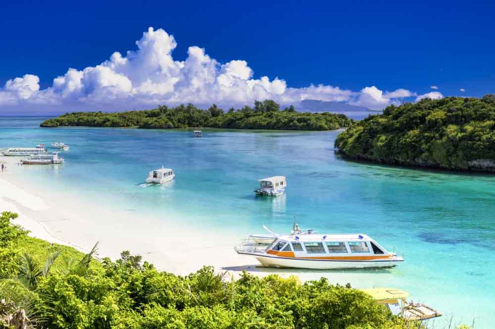 Tropical beach scene with clear turquoise water, white clouds, lush greenery, and small, anchored boats. The atmosphere is serene and inviting.