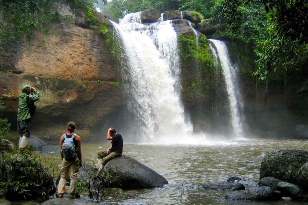 Three people viewing a powerful, misty waterfall cascading down a rocky cliff into a pool, set in lush greenery, conveying a sense of adventure.