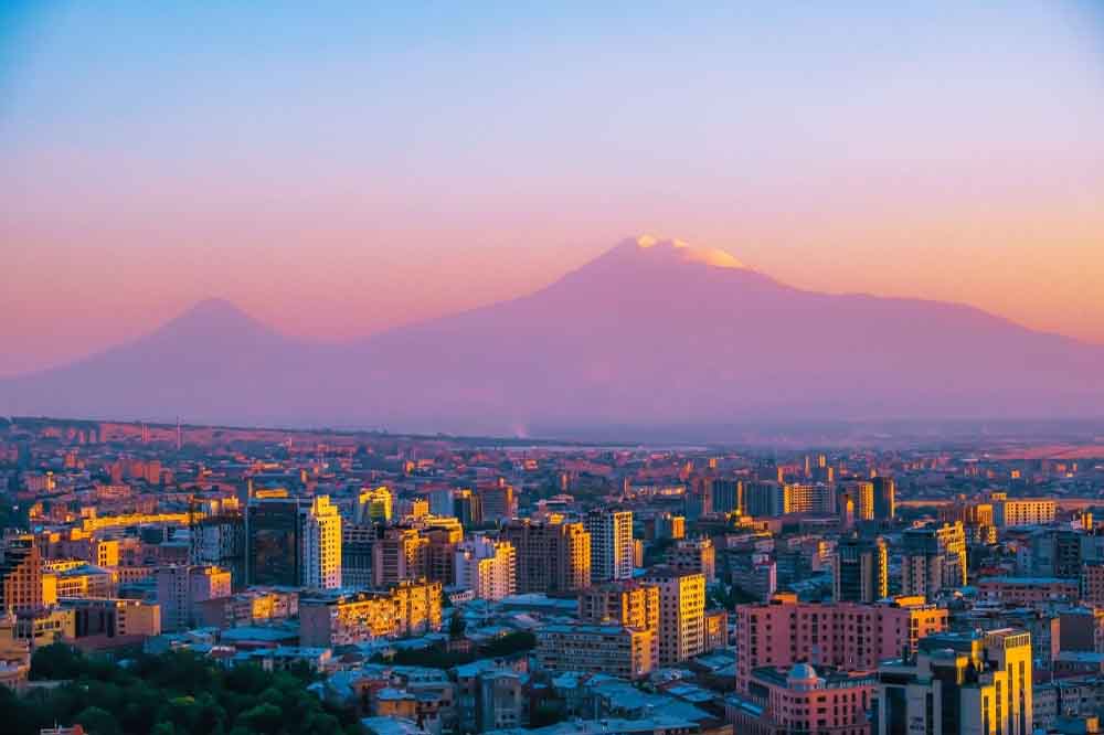Cityscape at sunset with orange-tinted buildings and distant snow-capped mountains against a soft pink and purple sky. Calm and serene atmosphere.