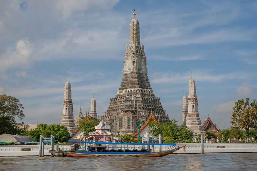 A vibrant long-tail boat floats on a river in front of the ornate, towering spires of the Wat Arun temple, against a clear blue sky in Bangkok, Thailand.