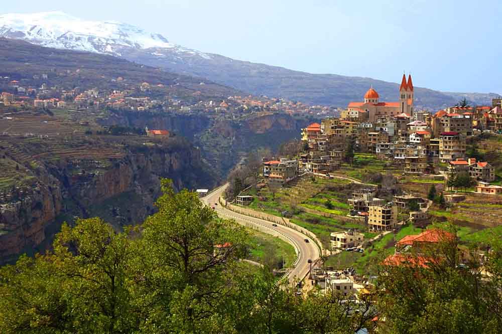A scenic hillside village with densely packed houses and a prominent church with red roofs. A winding road curves through green valleys under a snowy mountainous backdrop.