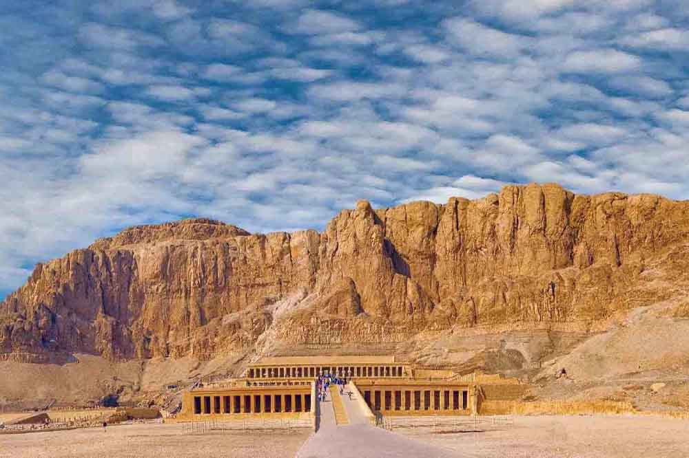 Ancient Egyptian temple nestled against rugged, sunlit cliffs under a partly cloudy blue sky. The scene is serene and majestic, highlighting history.
