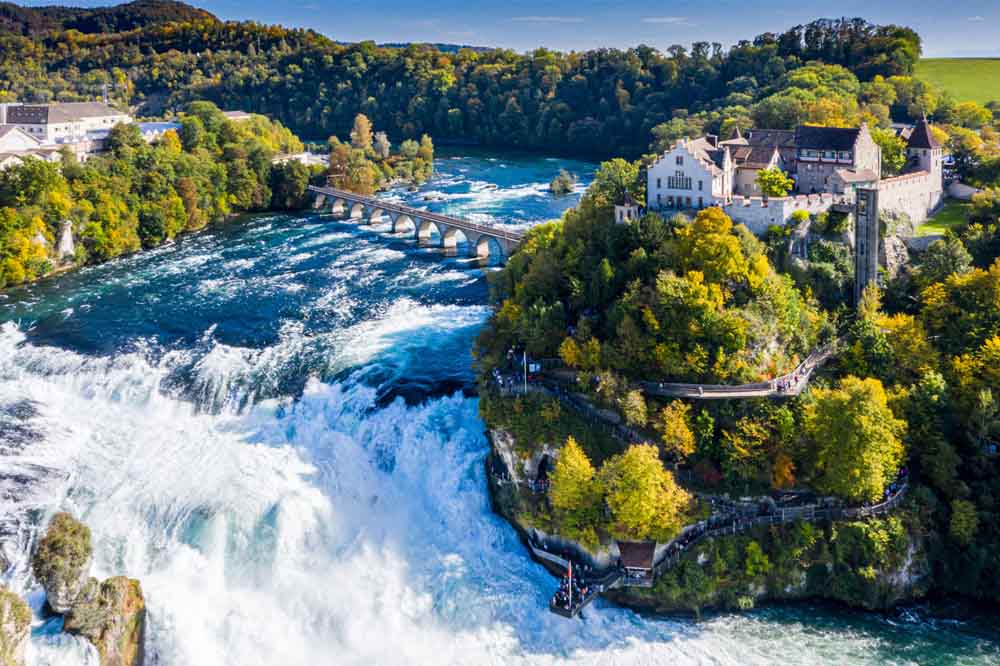 A stunning aerial view of the Rhine Falls in Switzerland, showcasing the powerful waterfall, a historic castle perched on a lush green hill, and a scenic arched bridge crossing the river — a popular highlight of Switzerland tour packages.