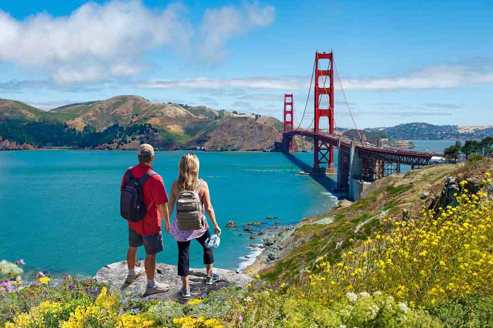 A couple with backpacks stands on a hillside overlooking the Golden Gate Bridge and bay. Bright flowers bloom in the foreground under a clear blue sky.