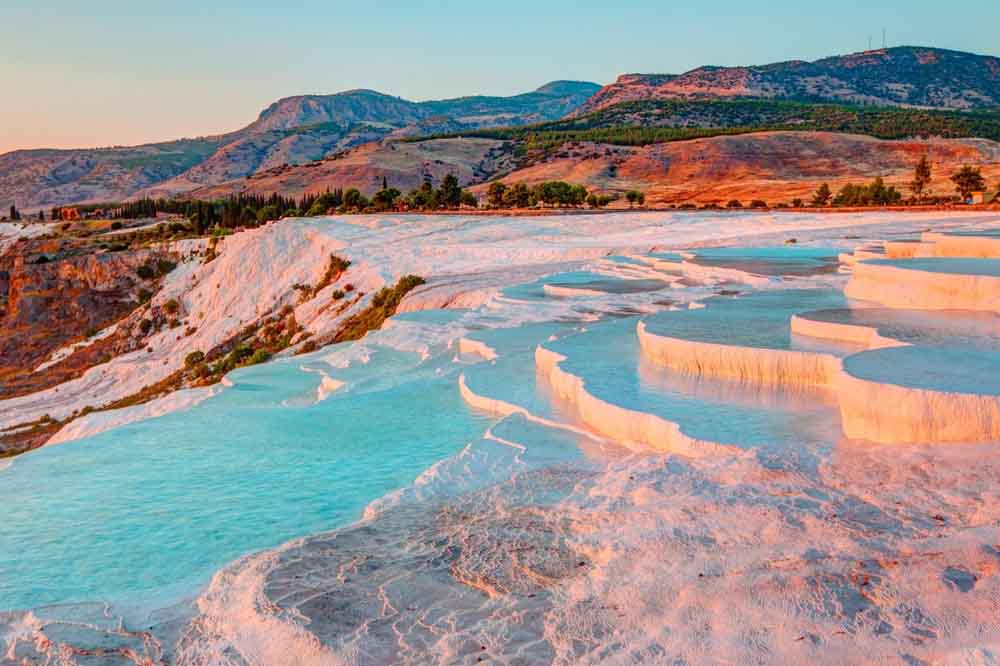 Sunlit terraced pools of blue water cascade over white travertine terraces at Pamukkale, Turkey, with a backdrop of green hills and mountains.