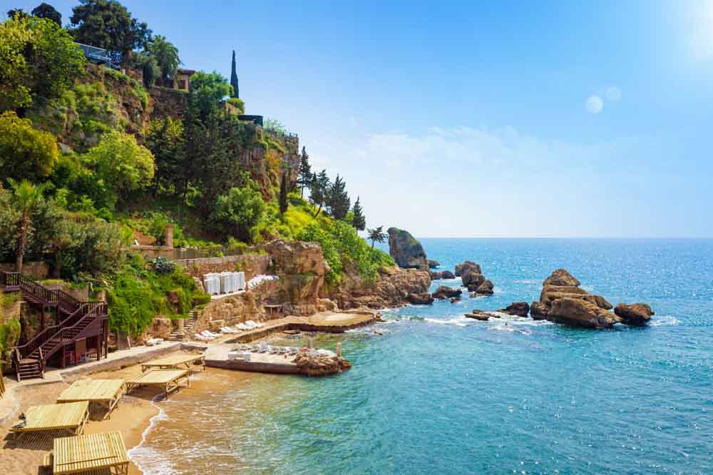 Rocky seaside cliff with lush greenery and a wooden staircase leading to a sandy beach. Sunlit blue ocean, exuding a serene, inviting atmosphere.