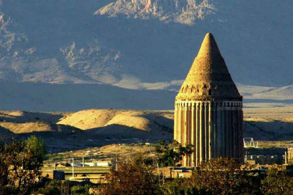 A tall, conical stone tower rises from a desert landscape, surrounded by small buildings and trees. Distant mountains provide a dramatic backdrop under a clear sky.