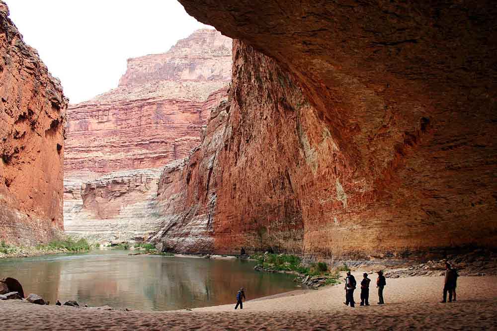 A group of people stand on sandy ground under a vast, rocky overhang, overlooking a calm river bordered by towering red canyon walls, evoking awe and tranquility.