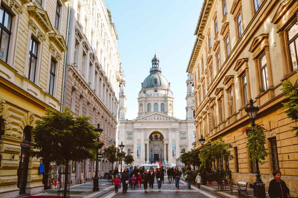 A bustling street lined with historic buildings leads to a grand cathedral with a large dome. People walk along, creating a lively, vibrant atmosphere.