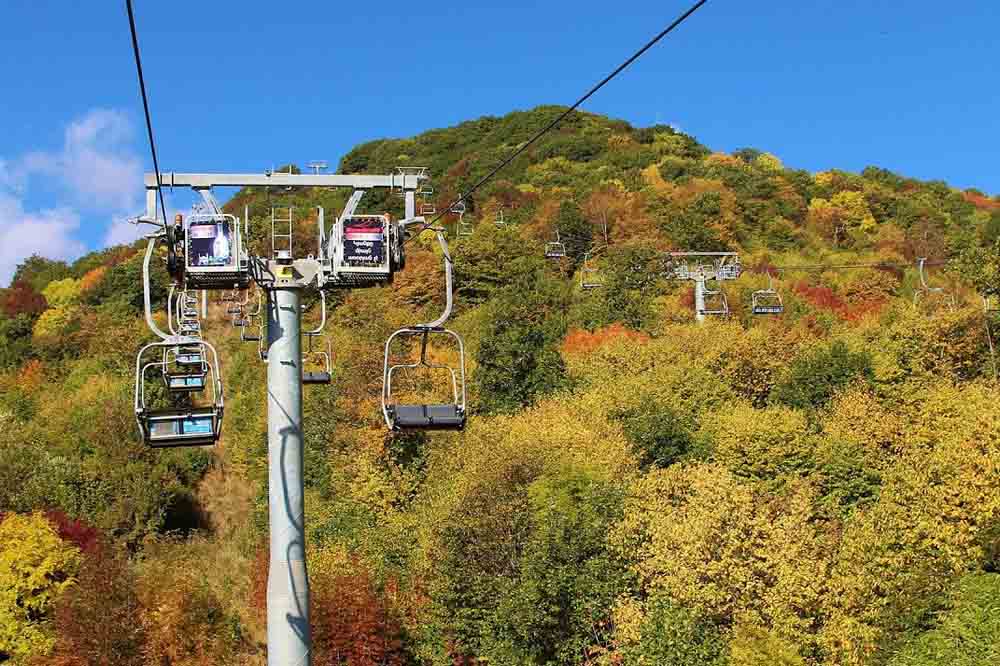 Chairlift ascending a hill covered in vibrant autumn foliage, with shades of green, yellow, and red, against a clear blue sky. Invokes a serene, picturesque mood.