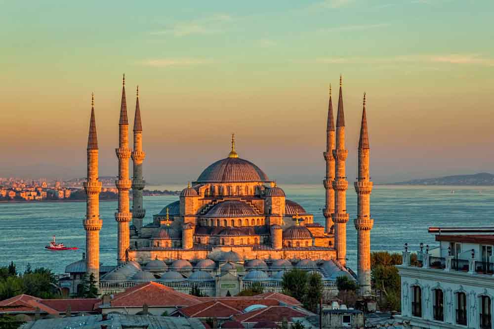 The Blue Mosque in Istanbul, illuminated by sunset light. Six minarets frame its domes, with a serene sea and distant cityscape in the background.
