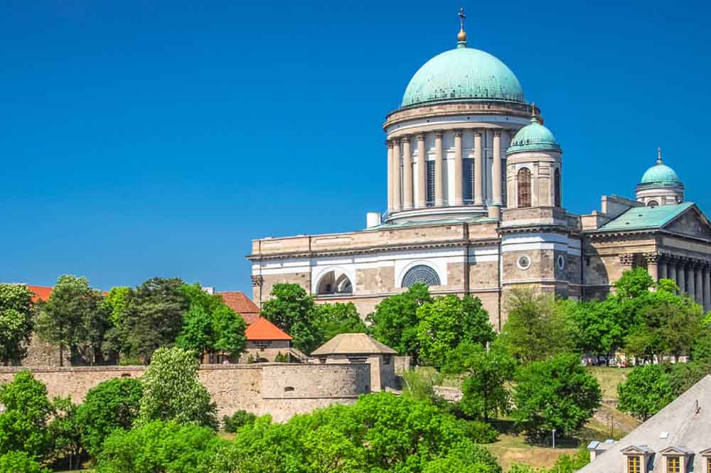 Majestic cathedral with a large green dome set against a clear blue sky; surrounded by lush trees and historic stone walls. Vibrant and serene.