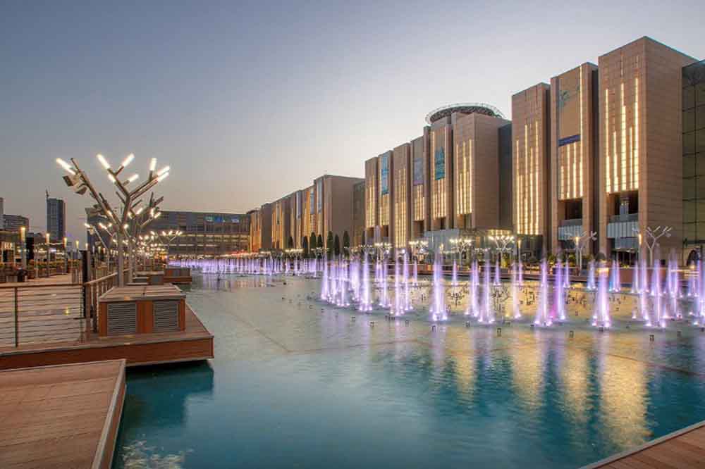 Modern architectural landscape at dusk with illuminated water fountains casting purple light in a reflective pool, flanked by a lit building facade.