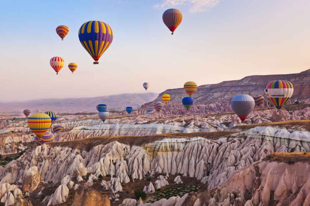 Colorful hot air balloons float above a rocky, canyon landscape at sunrise, creating a serene and vibrant scene against a pastel sky.