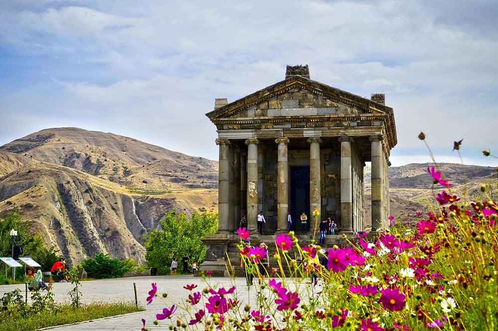 Ancient temple with columns sits against a mountainous backdrop, partly cloudy sky, and colorful wildflowers in the foreground, exuding a serene atmosphere.