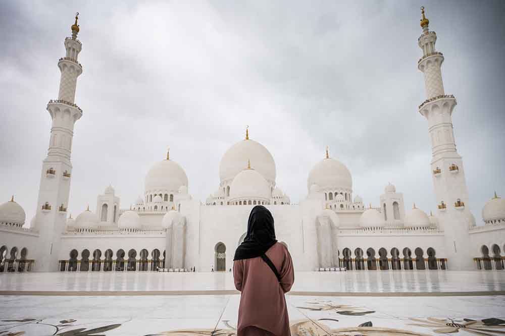 A person in dark hijab and pink coat gazes at the Sheikh Zayd Grand Mosque, under a cloudy sky, evoking a serene and contemplative mood.