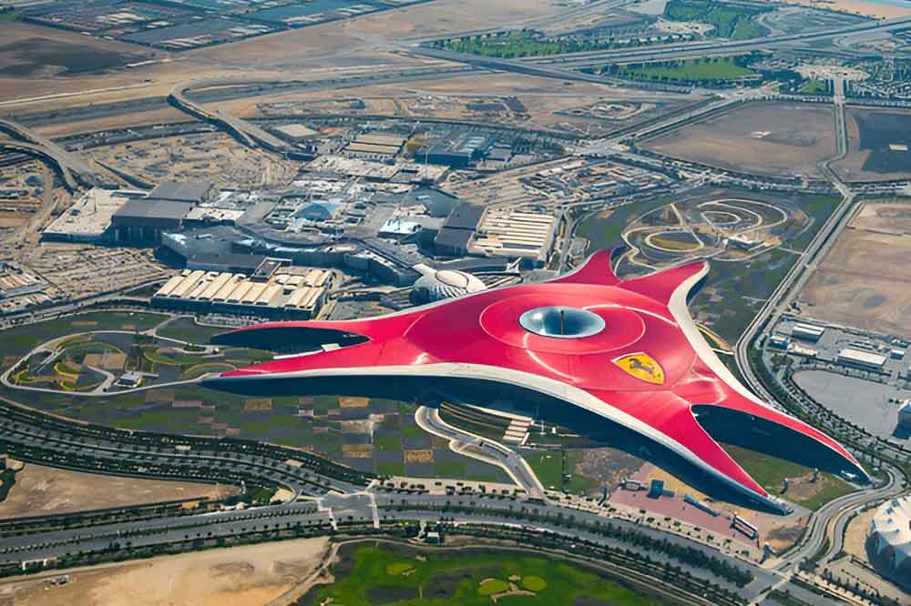Aerial view of a Ferrari world Dubai, red-roofed building shaped like a star, featuring the Ferrari logo. Surrounding it are roads, green spaces, and arid terrain.